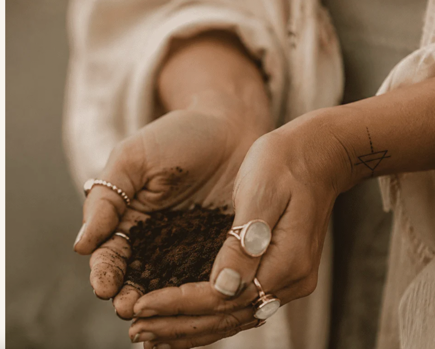 Hands with silver rings and a small wrist tattoo, gently holding dark soil