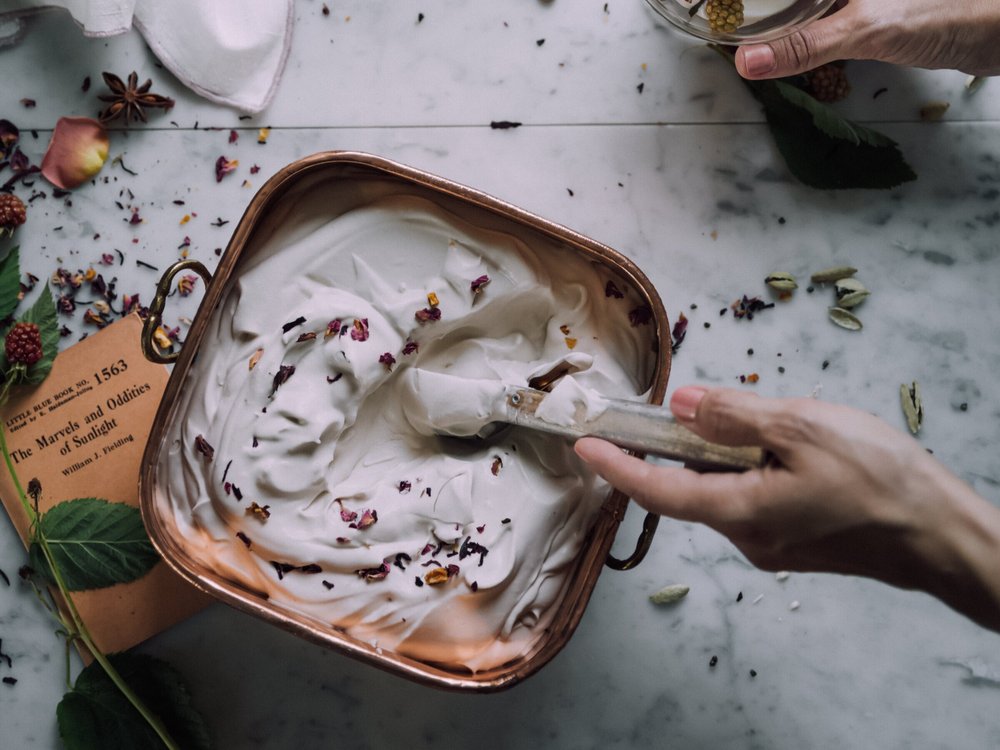 Botanical cream and rose petals in a copper bowl on marble, with fresh leaves and an old book